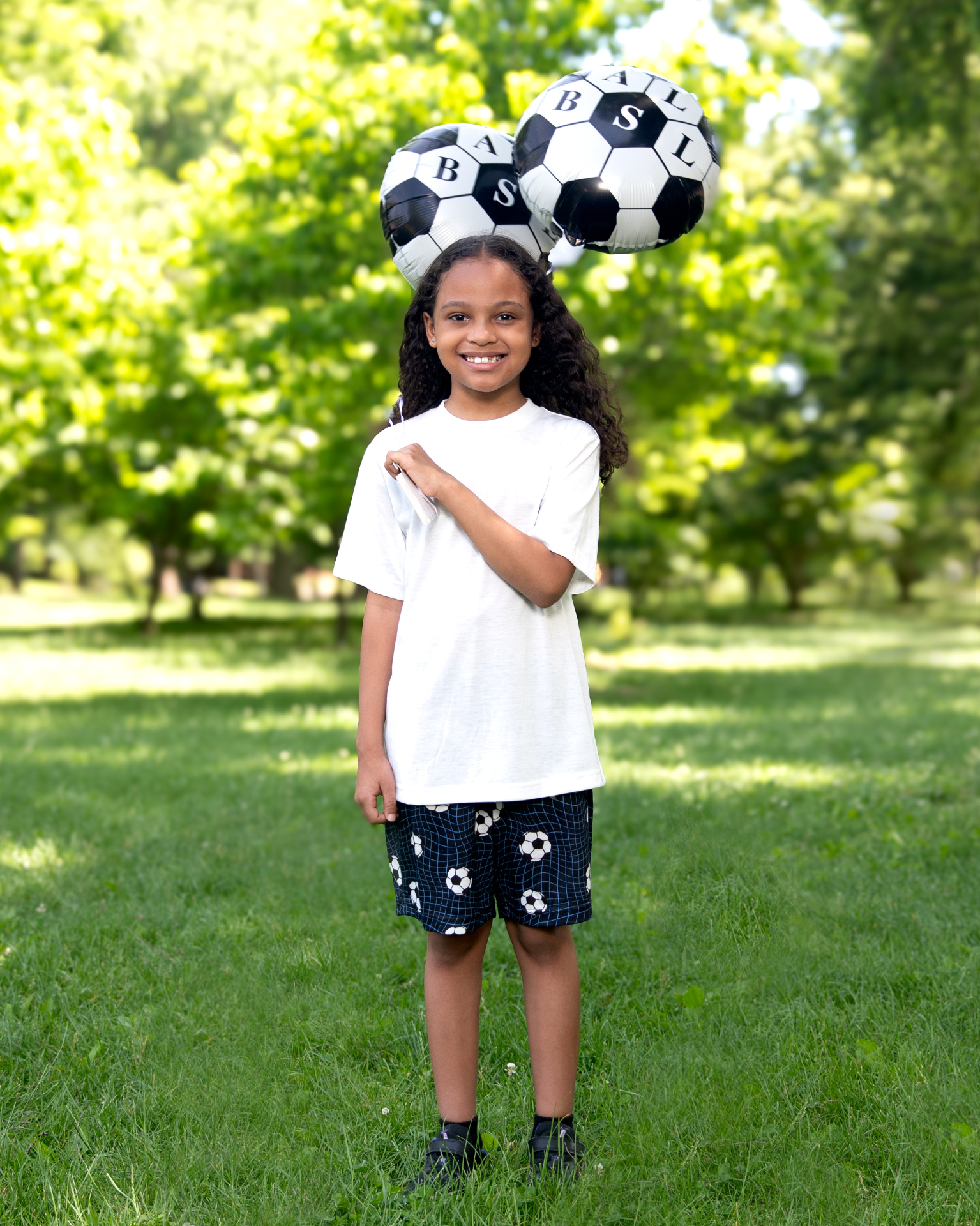 Bronx kid in a white tee holding two soccer-ball foil balloons in the park